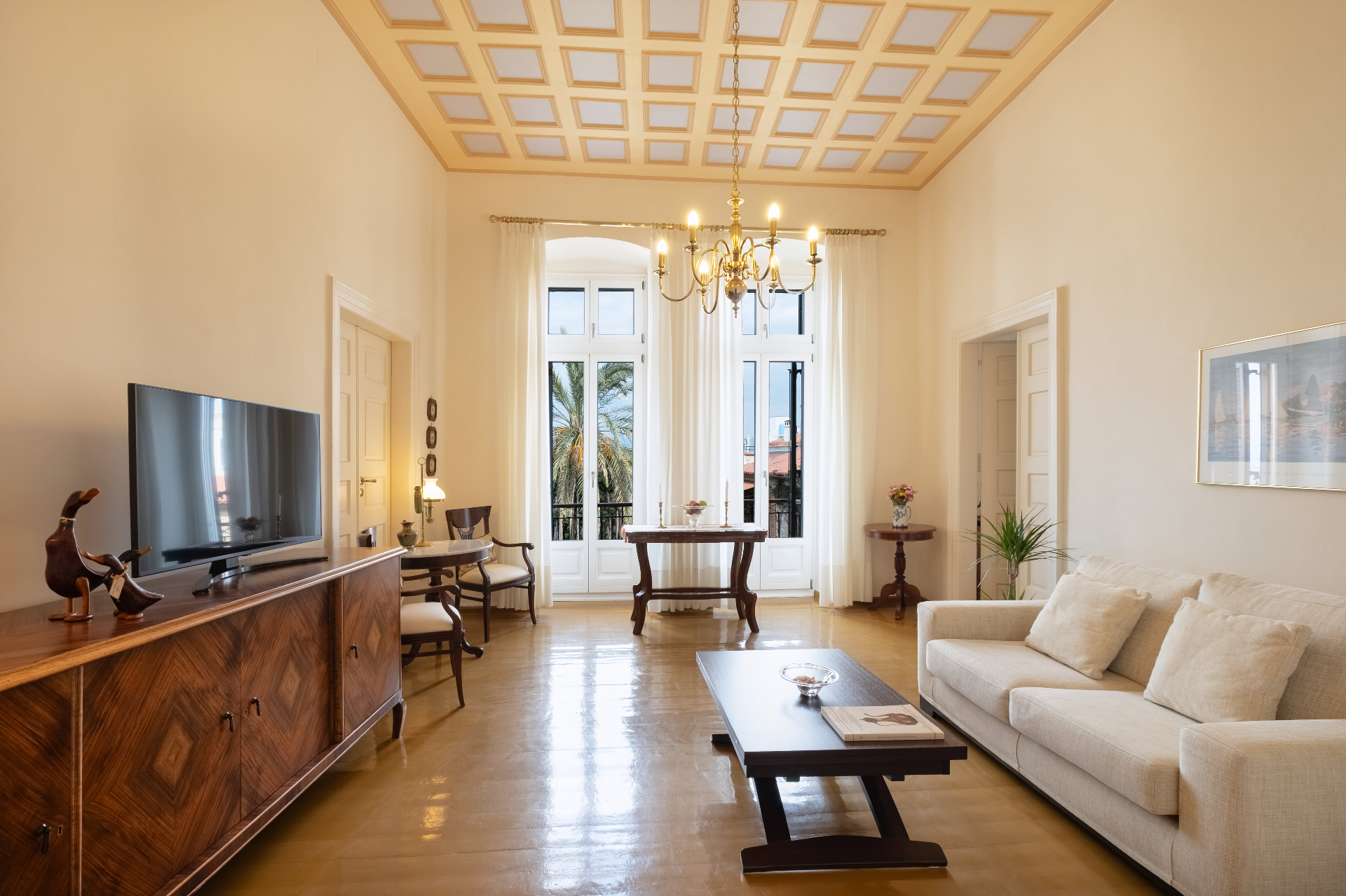 Main salon with painted coffered ceiling, chandelier, and tall balcony doors.